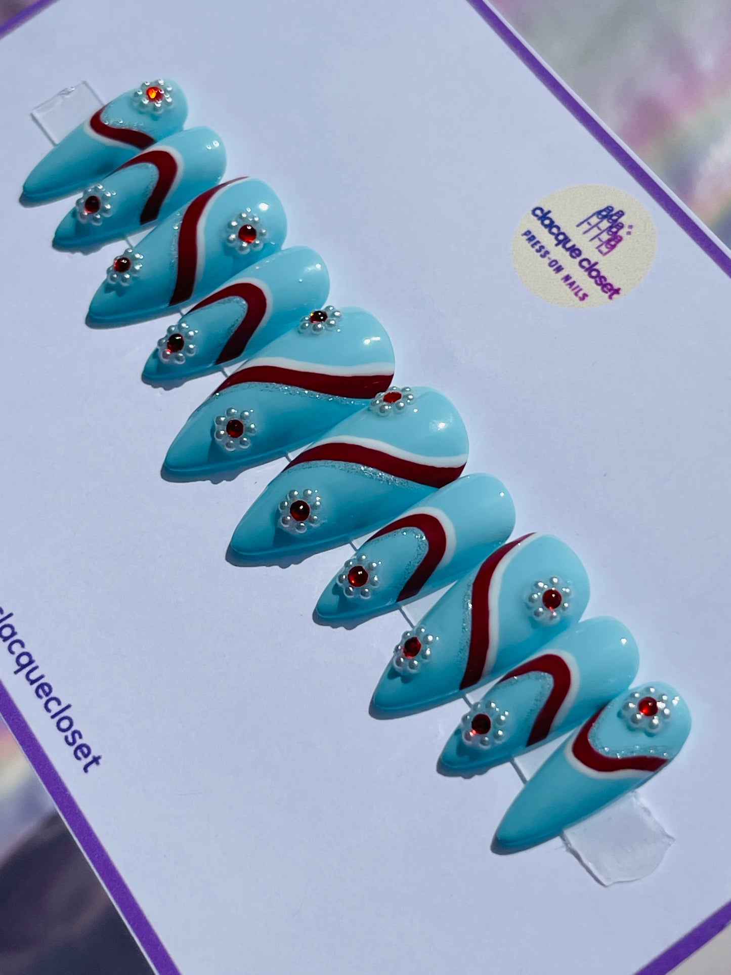 Medium-length stiletto nails in a vivid blue hue, featuring intricate designs with white, silver, and red stripes, and adorned with flowers made from pearls and red gems for a sophisticated touch.