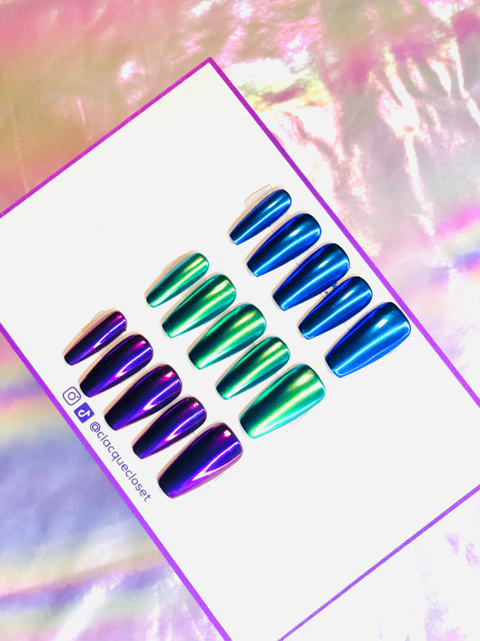 Three sets of chrome press-on nails displayed in a row, each with a lustrous metallic finish. The first set is in a vivid blue chrome, the second in a radiant green chrome, and the third in a deep purple chrome, each set shimmering and changing color subtly under different lighting.