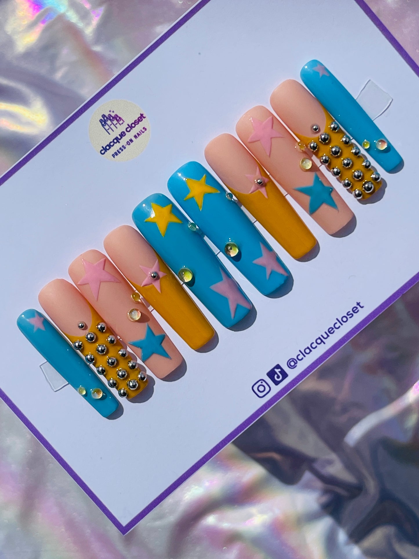 Long square nails painted with a celestial theme, showcasing blue, pink, and yellow stars scattered across a clear base, accented with tiny silver balls for added dimension.