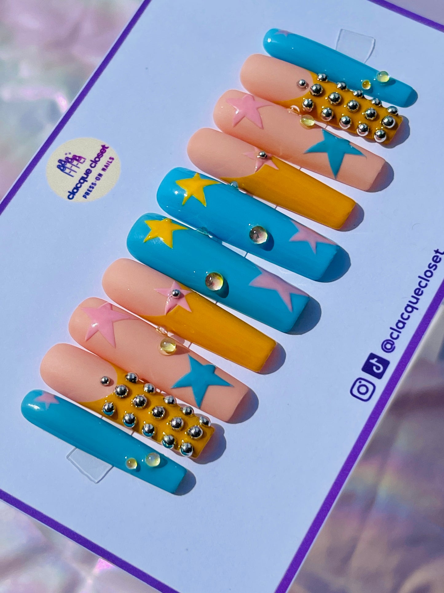 Long square nails painted with a celestial theme, showcasing blue, pink, and yellow stars scattered across a clear base, accented with tiny silver balls for added dimension.