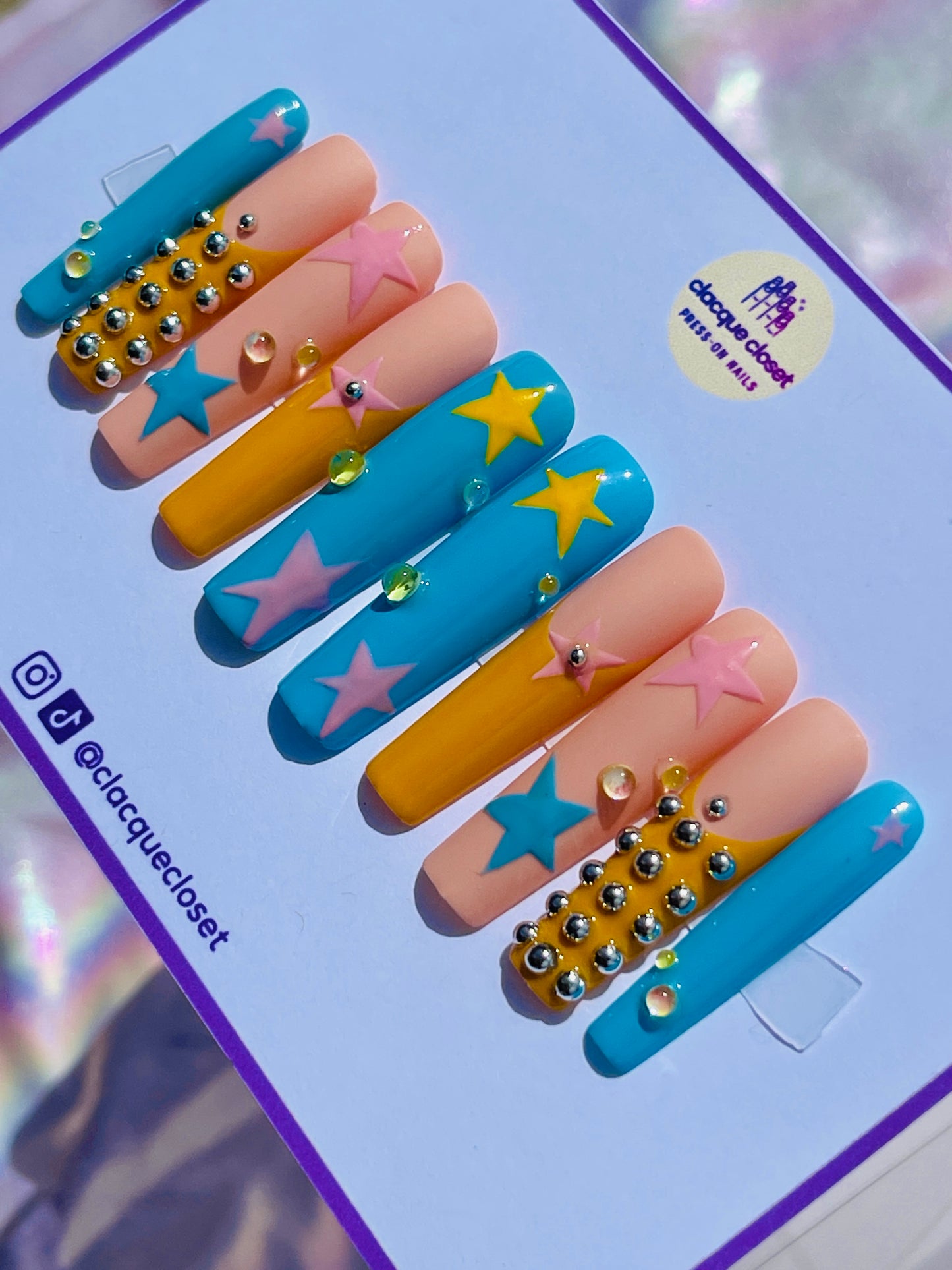 Long square nails painted with a celestial theme, showcasing blue, pink, and yellow stars scattered across a clear base, accented with tiny silver balls for added dimension.