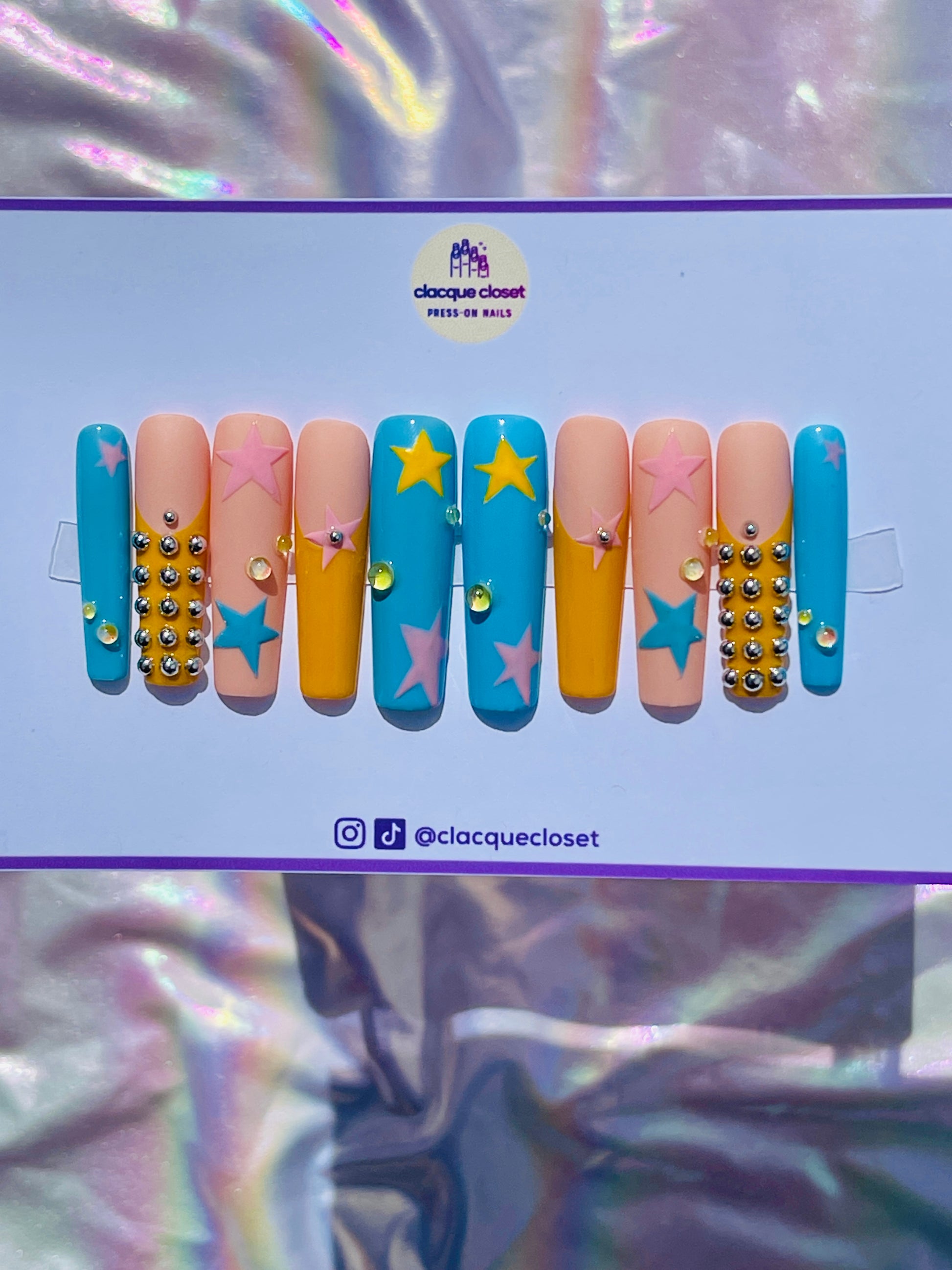 Long square nails painted with a celestial theme, showcasing blue, pink, and yellow stars scattered across a clear base, accented with tiny silver balls for added dimension.