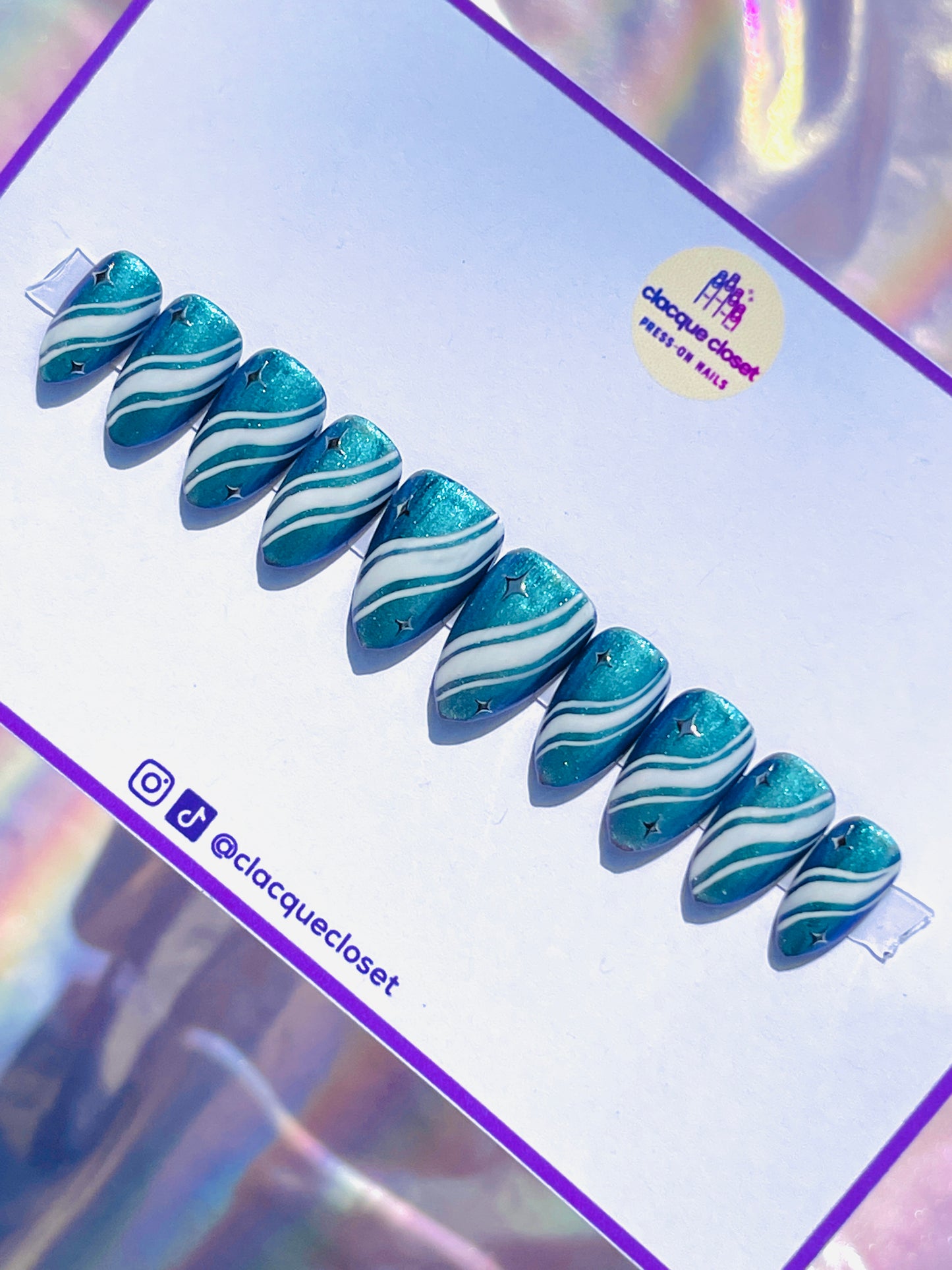 Medium-length almond-shaped nails in a sparkly blue color, detailed with white curvy lines and adorned with silver stars, creating a whimsical night sky effect.
