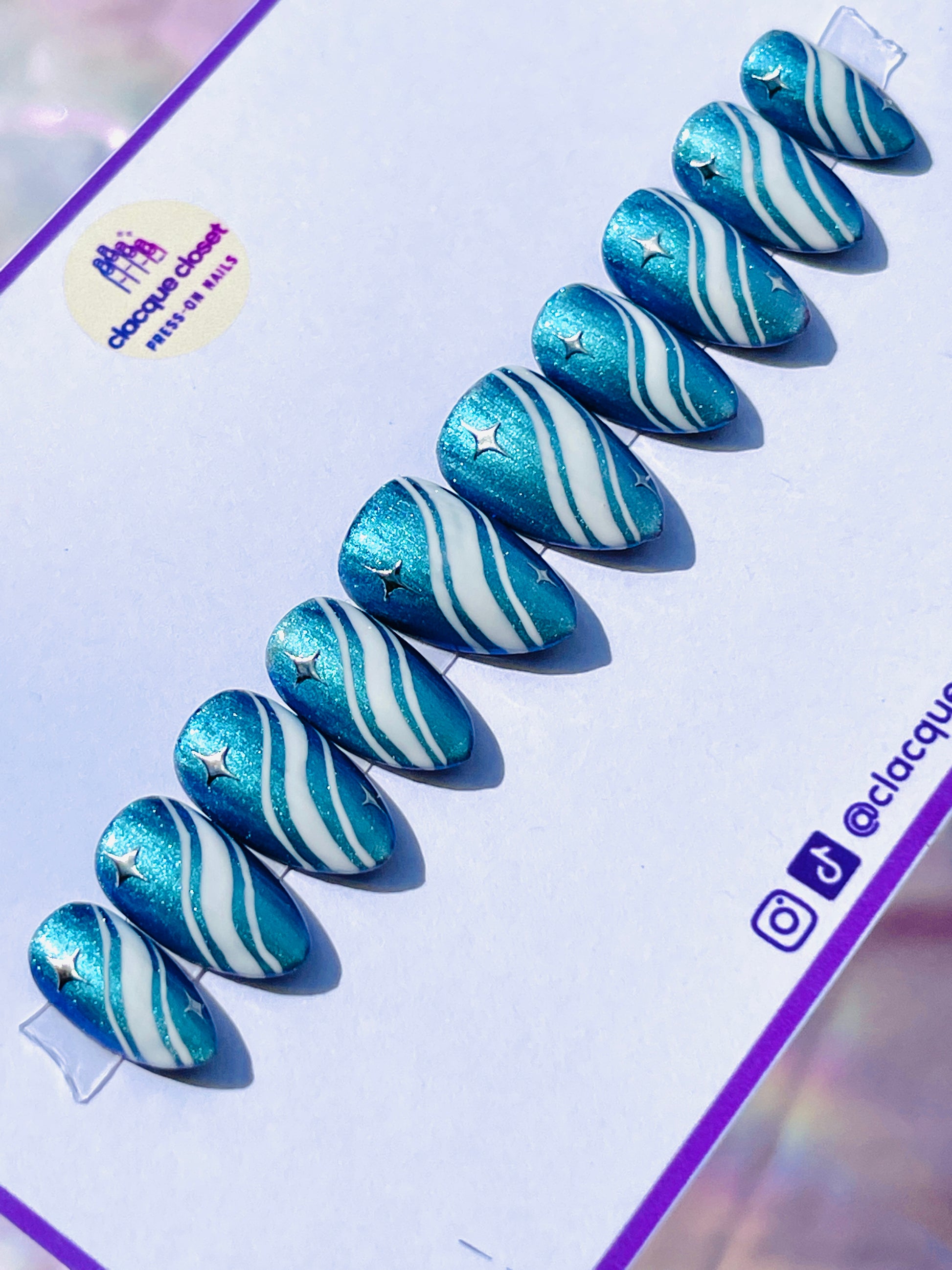 Medium-length almond-shaped nails in a sparkly blue color, detailed with white curvy lines and adorned with silver stars, creating a whimsical night sky effect.