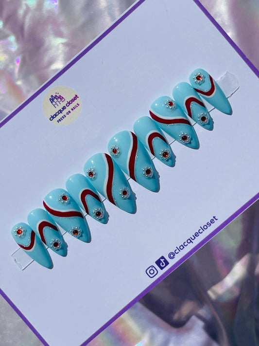Medium-length stiletto nails in a vivid blue hue, featuring intricate designs with white, silver, and red stripes, and adorned with flowers made from pearls and red gems for a sophisticated touch.