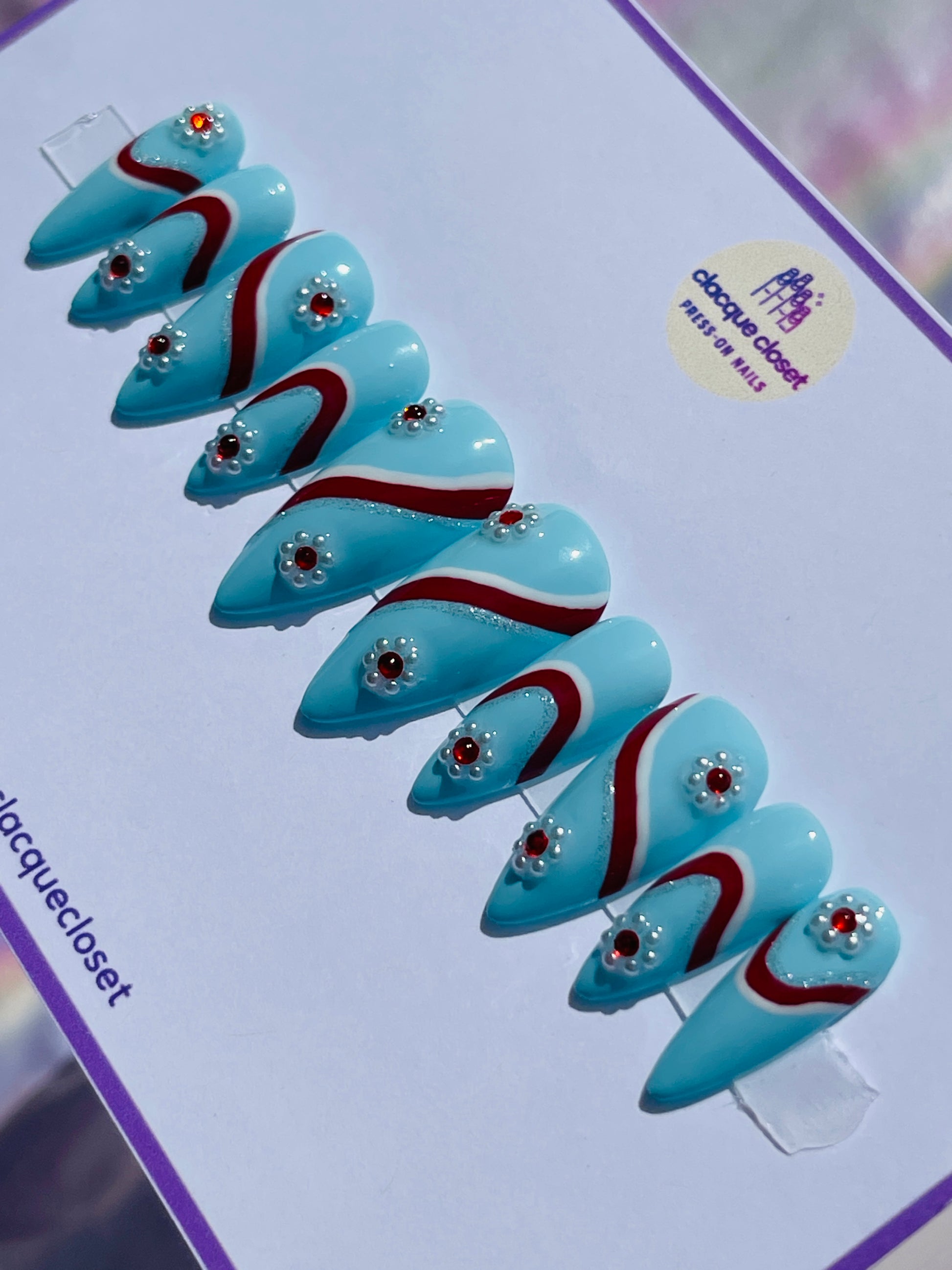 Medium-length stiletto nails in a vivid blue hue, featuring intricate designs with white, silver, and red stripes, and adorned with flowers made from pearls and red gems for a sophisticated touch.